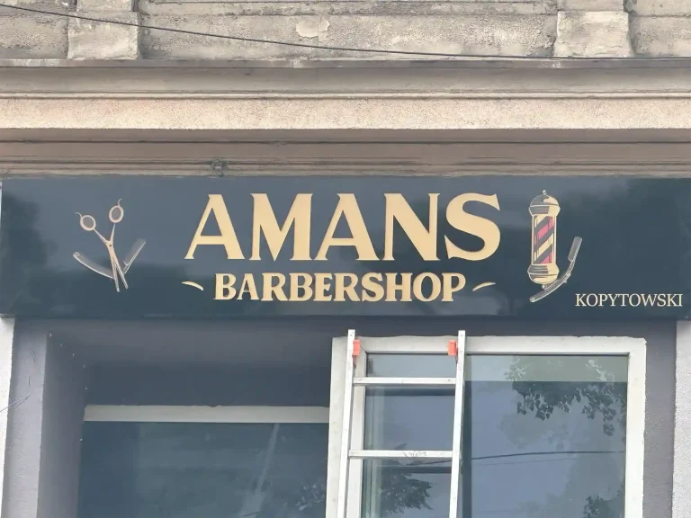 Schwarzes Firmenschild mit goldener Beschriftung fuer Amans Barbershop an der Fassade